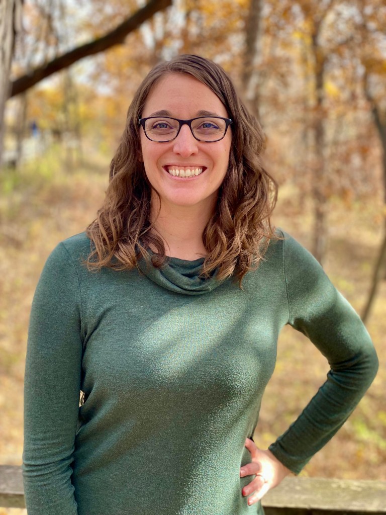 A middle-aged woman with shoulder-length curly hair is wearing plastic-framed glasses and a dark green sweater. She is smiling, has her hand on her hip, and is among some trees displaying autumn colors. 